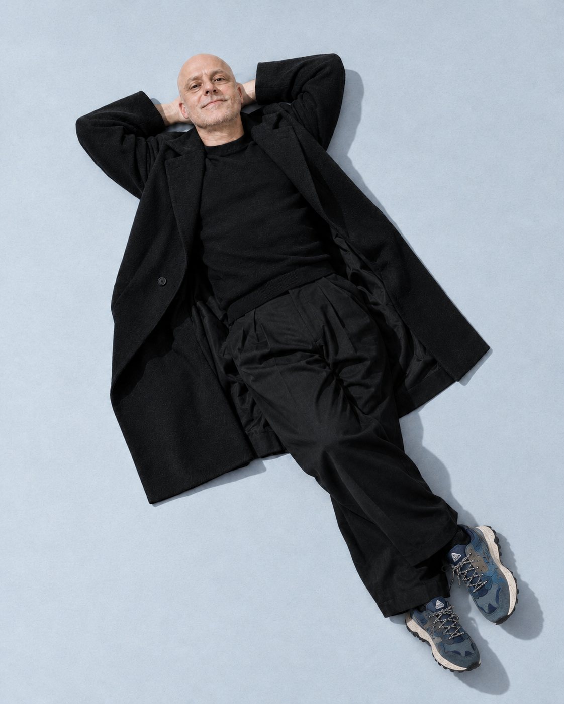 Julien Vallet allongé sur un papier bleu, manteau noir, baskets bleues, les mains derrière la tête.